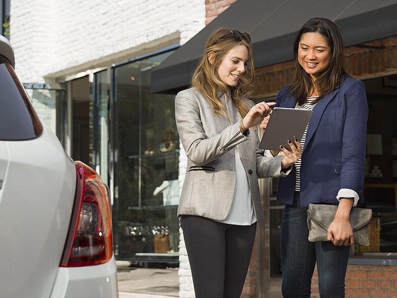 Two women looking at a tablet next to a new car.