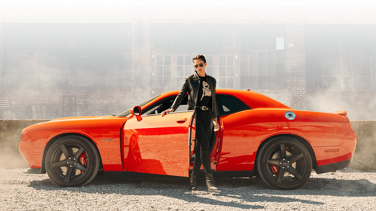 Woman stepping out of her orange 2018 Dodge Challenger amid a cloud of dust.