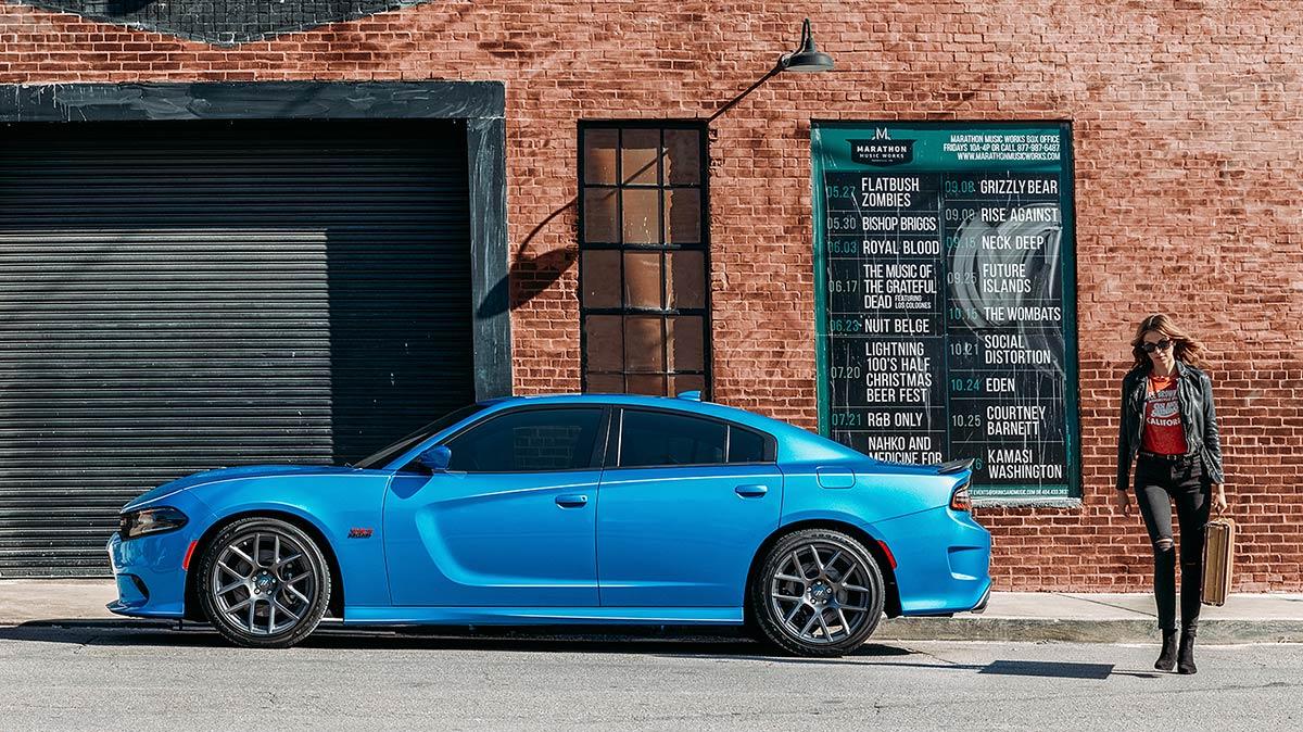 A woman walking away from her blue 2018 Challenger parked next to a brick building.