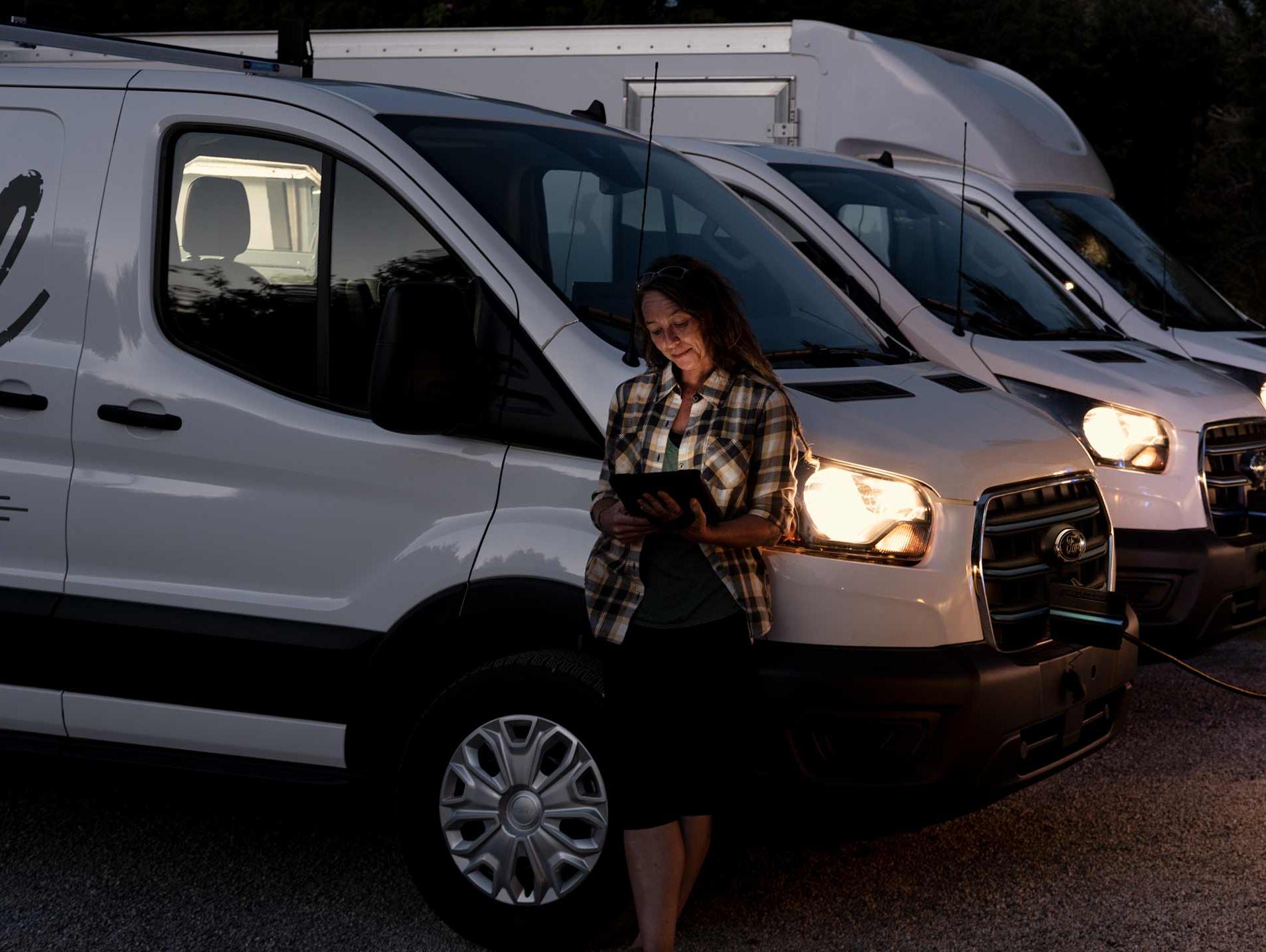 A row of 2024 E-Transits at night charging up with a woman leaning against one of them