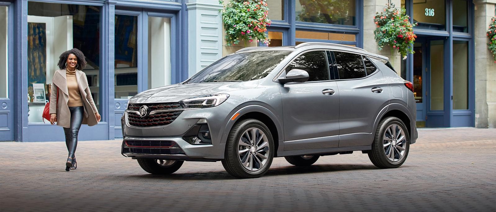 A 2023 Encore GX Essence in Moonstone Gray Metallic parked on a street of retail shops with a woman walking by.