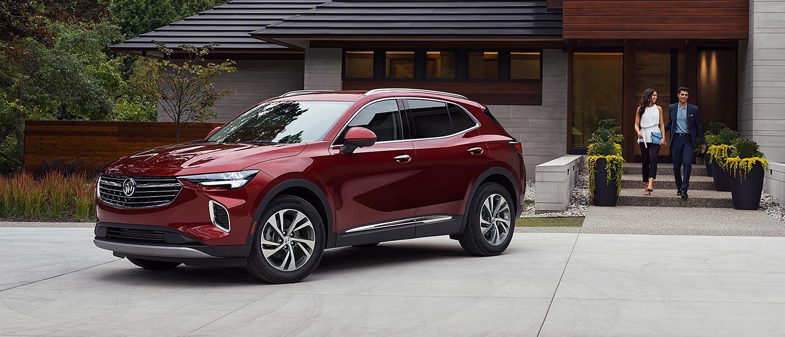 A 2023 Envision Essence AWD in Cinnabar Metallic red parked in driveway in front of house with a couple approaching.