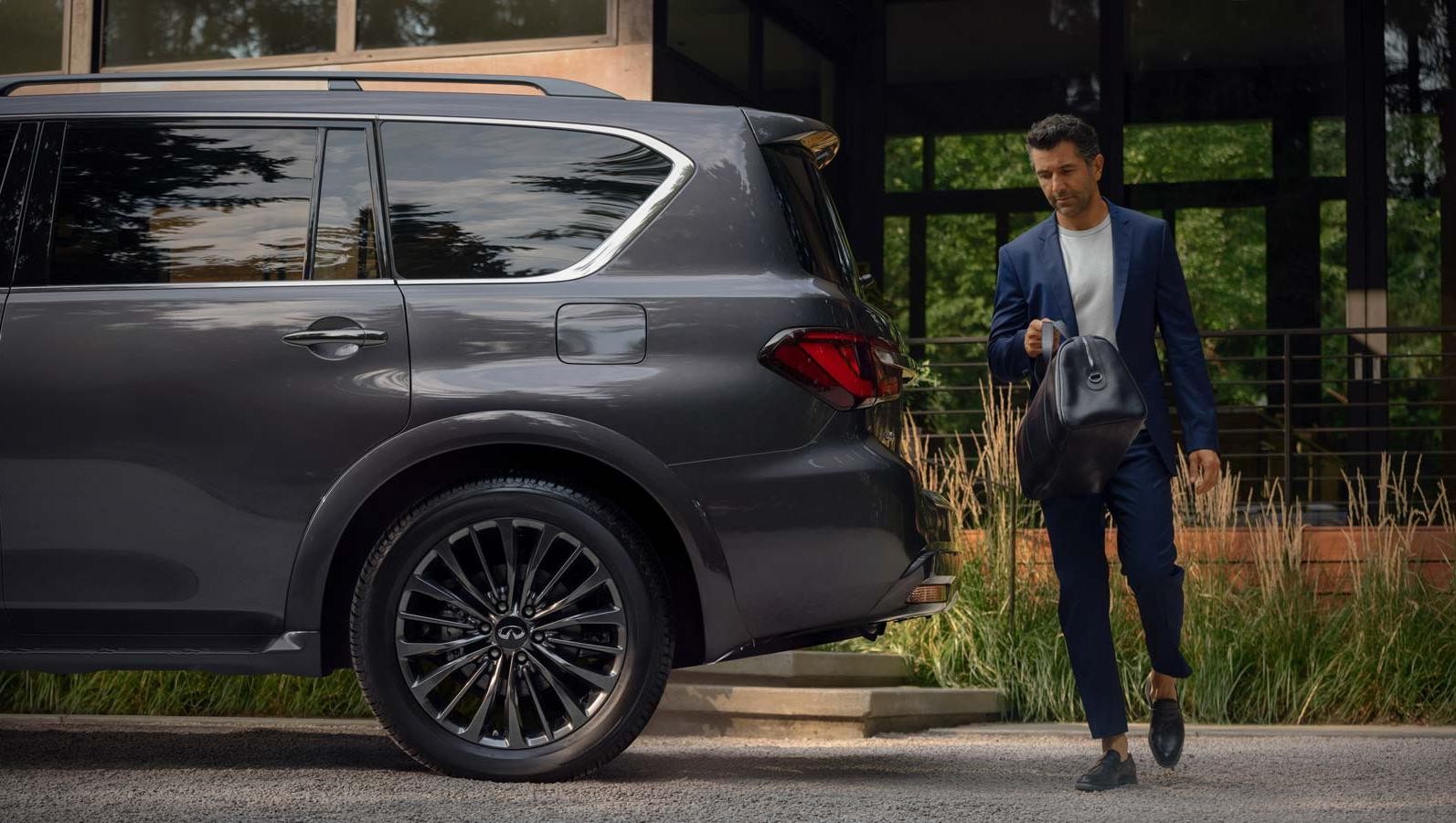 Man dressed in suit walking behind a 2023 QX80 with a suitcase