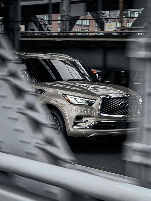 A peek-a-boo view of a grey INFINITI QX80 driving over a bridge.