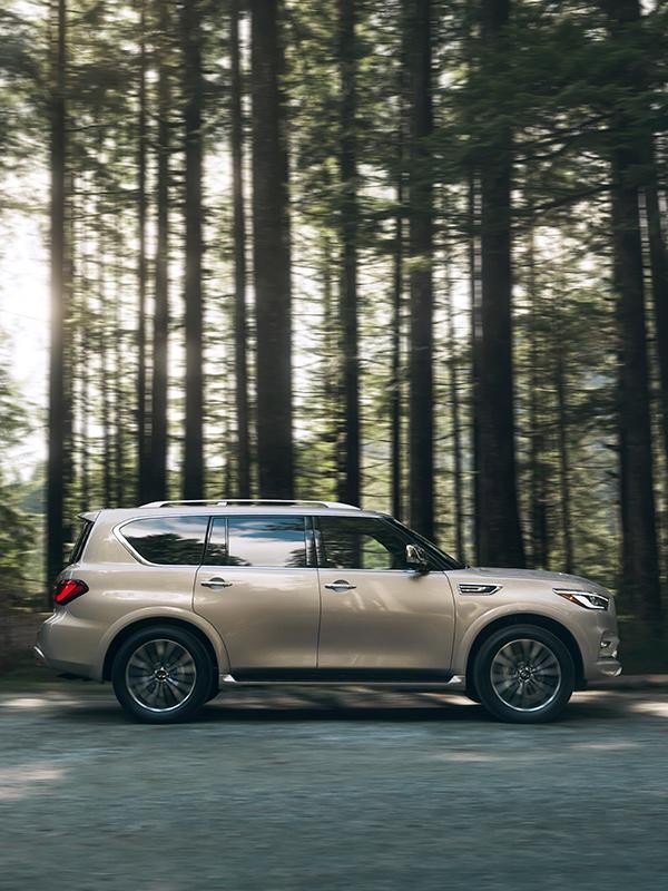 A profile view of a tan INFINITI QX80 on a forest road.