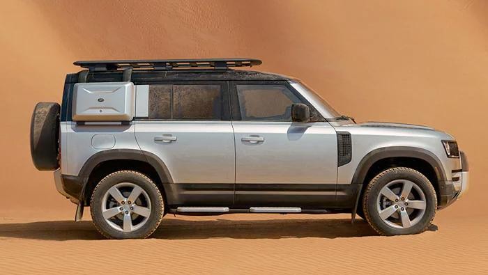 Profile of a silver Land Rover Defender in front of a sand dune.