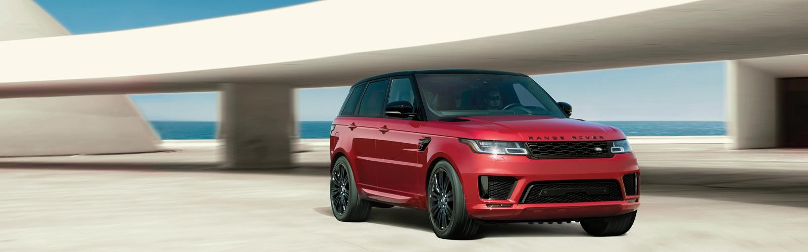 Front corner view of a red Range Rover Sport parked next to an ocean-side overpass.