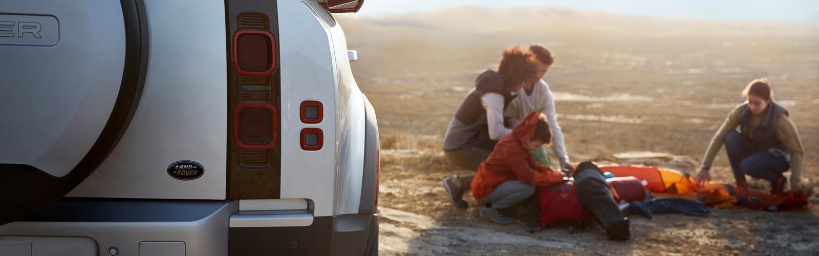 Close-up view of a Land Rover Defender rear corner with a family setting up a campsite in the background. 