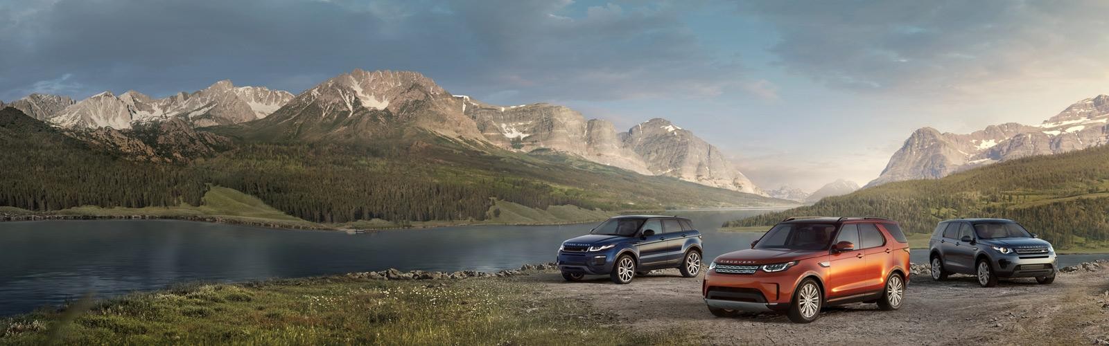 Three Land Rover models parked at a scenic spot in front of a lake and mountain range.