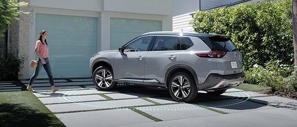 A woman walking towards her grey Nissan Rogue parked in the driveway.