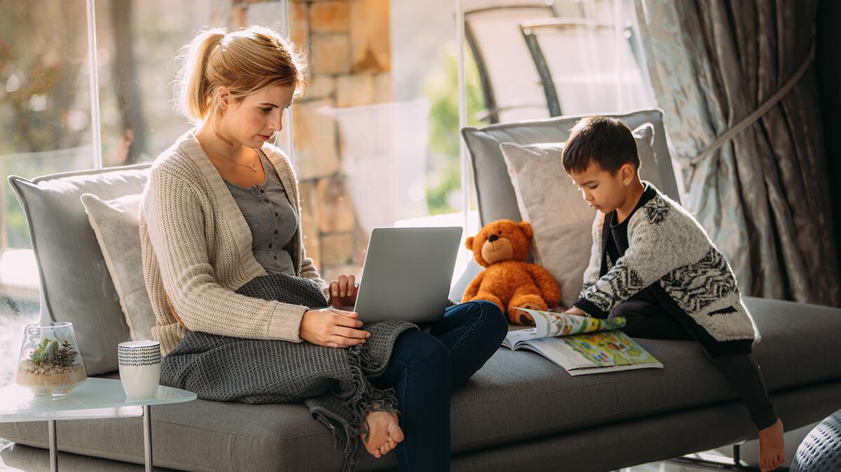 Woman using a laptop at home while a young boy plays beside her