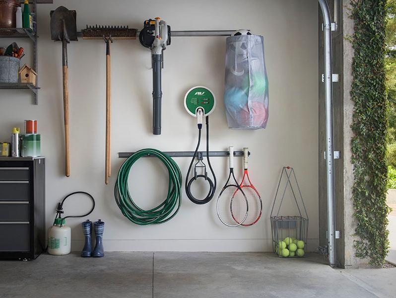 An EV charging port setup in a home garage mounted on the wall next to hanging garden tools and sports equipment.
