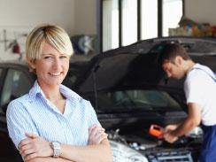 woman with arms folded smiling in front of a car with hood open, service tech working on car