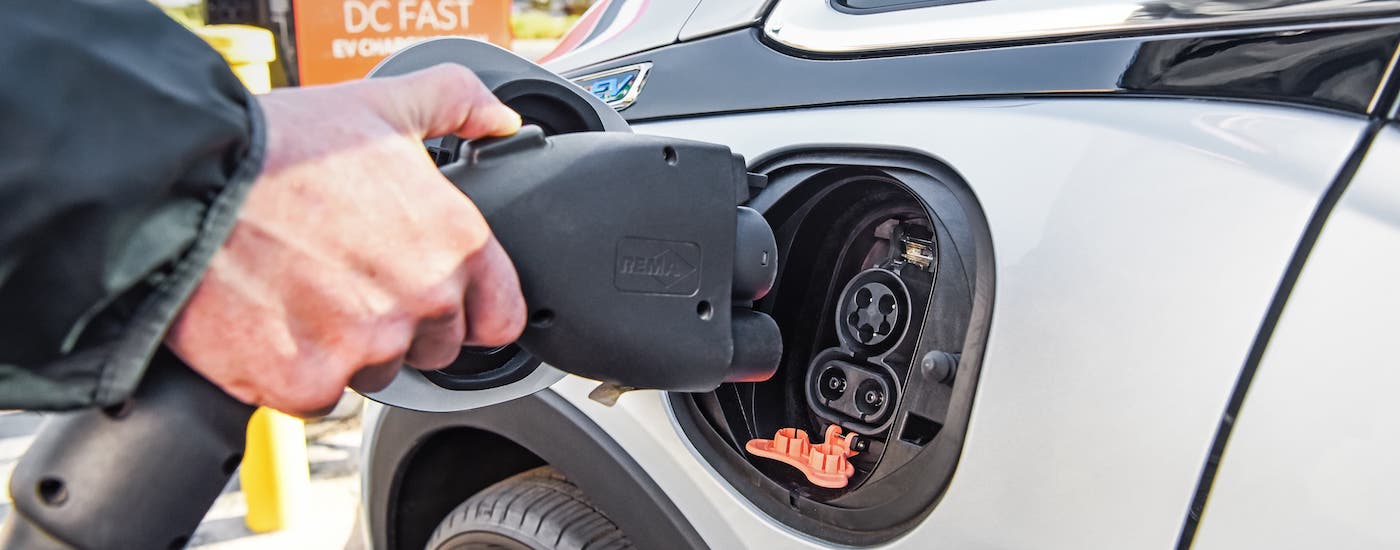 A closeup shows a hand plugging in a silver 2018 Chevy Bolt EV.